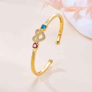 May include: A gold-toned cuff bracelet featuring a central heart motif encrusted with small clear stones. The bracelet includes two round-cut stones in blue and red, with the engraved names George and Caroline positioned near each stone.