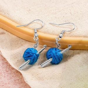 Peut inclure: Une paire de boucles d'oreilles à crochet argentées représentant une miniature de pelote de laine bleue traversée par un petit crochet argenté. Les boucles d'oreilles sont suspendues à des attaches standard, illustrant les arts du fil.