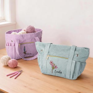 May include: Two corduroy craft bags in lavender and light blue, each featuring a zippered front pocket, side slots for tools, and embroidered floral designs with the names Olivia and Emily. Three pink crochet hooks and two yarn balls are nearby.