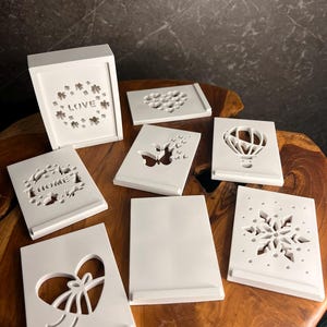 Könnte beinhalten: Eine Sammlung weißer Dekorationsartikel mit ausgeschnittenen Designs, darunter eine Box mit Kleeblattmuster und dem Wort "LOVE" sowie mehrere rechteckige Ständer mit Herz-, Schmetterlings-, Heißluftballon-, Schneeflocken- und "HOME"-Designs.
