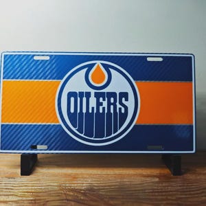 May include: A rectangular license plate featuring the Edmonton Oilers logo. The plate has a blue carbon fiber pattern background with orange stripes and the team's logo in the center. The word "OILERS" is prominently displayed.