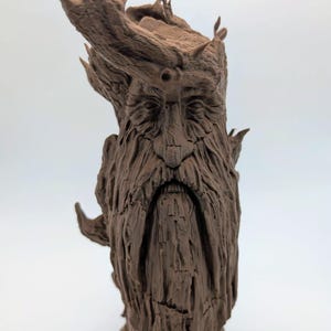May include: A brown, detailed sculpture of a bearded face with a wooden texture. The sculpture has a long beard, furrowed brow, and leafy accents on the head, resembling a tree spirit or mythical figure. The sculpture is set against a white background.