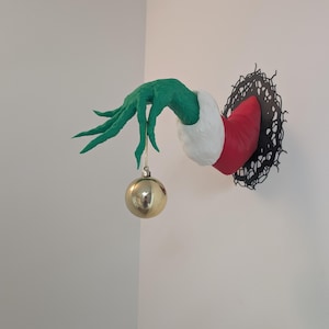 May include: A wall-mounted sculpture featuring a textured green hand emerging from a black, jagged, circular frame. The hand wears a red sleeve with a white cuff and holds a single, smooth, reflective gold spherical ornament by a thin string.