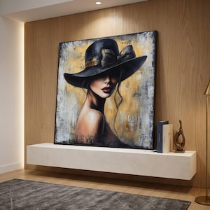 Puede incluir: Una obra de arte enmarcada que representa a una mujer con un gran sombrero negro con un lazo dorado. El fondo es abstracto, dorado y blanco. La mujer lleva pintalabios rojo y mira por encima del hombro. La obra está sobre una estantería blanca.