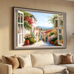 May include: A framed canvas print showing an Italian street scene through an open window. The artwork features white buildings, red-tiled roofs, and vibrant flowers in pots, with a cobbled street leading to distant mountains.