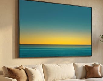 May include: Large framed wall art featuring a seascape with a gradient of colors. The artwork displays a blue sky transitioning to a yellow horizon over turquoise water. The frame is black, and the artwork is displayed above a cream-colored sofa with throw pillows.