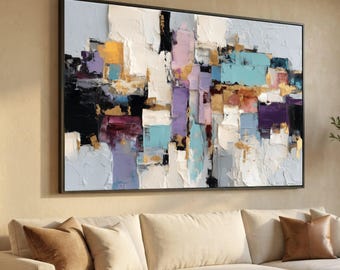 May include: Abstract painting with a black frame. The artwork features a textured surface with a palette of colors including white, black, gold, purple, and teal. The painting is displayed above a cream-colored sofa.