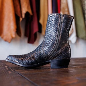 Könnte beinhalten: Ein schwarzer und brauner Lederstiefel mit Schlangenmuster. Der Stiefel hat einen seitlichen Reißverschluss und einen niedrigen Absatz. Der Stiefel wird auf einer Holzoberfläche mit Lederproben im Hintergrund präsentiert.