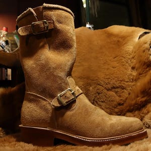 May include: A tan suede mid-calf boot featuring a rounded toe, a stacked heel, and two adjustable straps with metallic buckles. One buckle is positioned across the ankle area, while the second buckle is located at the top of the boot shaft.
