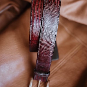 Puede incluir: Un cinturón de color burdeos oscuro con un patrón texturizado de piel de lagarto en toda la tira. El cinturón incluye una trabilla de cuero a juego y una hebilla de metal plateado pulido con un solo perno, con costuras detalladas en los bordes.
