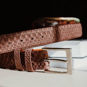 Puede incluir: Un cinturón marrón con un patrón texturizado de piel de serpiente y una hebilla de metal plateado. El cinturón cuenta con dos trabillas a juego y una serie de agujeros perforados para el ajuste, mostrando una textura detallada de escamas.