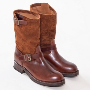Könnte beinhalten: Ein Paar wadenhohe Stiefel mit einem unteren Teil aus glattem braunem Leder und einem oberen Schaft aus strukturiertem braunem Wildleder. Jeder Stiefel verfügt über einen dekorativen Lederriemen über dem Spann mit Metallschnalle und eine kleine Schnalle.