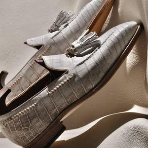 Puede incluir: Un par de mocasines blancos con un patrón texturizado de imitación de cocodrilo, punteras puntiagudas y borlas decorativas en el empeine. Los zapatos tienen un tacón bajo de madera, un borde de cuero trenzado y un forro interior marrón oscuro.