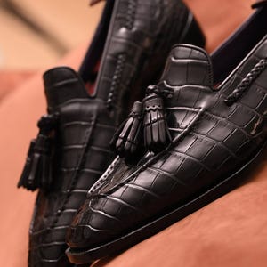 Puede incluir: Un par de mocasines negros con una textura brillante de relieve de cocodrilo. Cada zapato está adornado con una borla decorativa en el empeine y un borde de cuero trenzado a lo largo del cuello. Los zapatos tienen punta afilada y suela oscura.