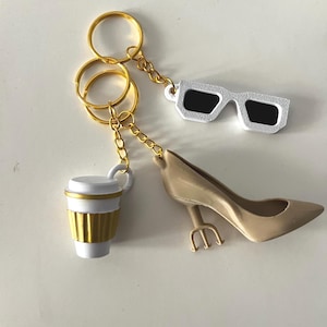 Può includere: Portachiavi con una tazza da caffè bianca con dettagli dorati, un paio di occhiali da sole bianchi con lenti nere e una scarpa con tacco beige con tacco a tridente. Tutti i portachiavi hanno anelli e catene dorate.