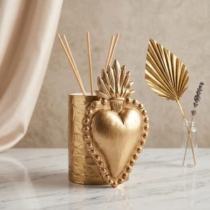 Puede incluir: Un conjunto decorativo dorado con un recipiente cilíndrico texturizado con palitos difusores, un adorno en forma de corazón y una hoja dorada. El adorno de corazón tiene un borde con cuentas y una parte superior en forma de llama.