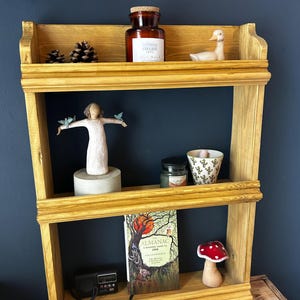 May include: A three-tiered wooden shelf unit with decorative items. The top shelf has a glass jar, pine cones, and a duck figurine. The middle shelf displays a figurine, a candle, and a patterned cup. The bottom shelf has a book, a radio, and a mushroom figurine.