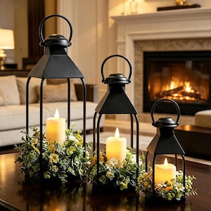 May include: Three black metal lanterns of varying heights, each featuring a top handle and a pyramid-shaped roof. Inside each lantern sits a cream-colored pillar candle surrounded by a base of green foliage and small white and yellow flowers.