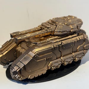 May include: A bronze-coloured model of a futuristic tank. The tank has a dual-barrelled turret and detailed paneling, mounted on a black base. The design suggests a science fiction or military theme.