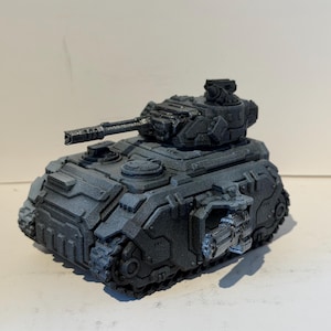 May include: A dark grey toy tank with a detailed design, featuring a large turret and visible tracks. The miniature tank has a textured surface. The model is set against a plain background.