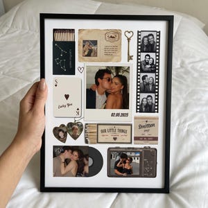 Op de afbeelding: Een ingelijste collage met een zwart luciferdoosje met sterrenbeelden, een vintage ansichtkaart, een gouden sleutel, een filmstrip met foto's, een speelkaart met de tekst Lucky You, hartvormige fotobedels, een luciferdoosje en een camera.