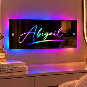 以下が含まれることがあります： 黒いアクリル製の長方形の壁掛け看板。中央には「Abigail」という名前が光るマルチカラーの筆記体で描かれています。看板の背面からは鮮やかな紫、青、ピンクのLEDライトが照射されており、銀色の金具で固定されています。