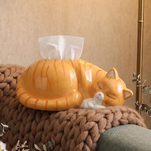 Puede incluir: Caja de pañuelos de cerámica con forma de gato atigrado naranja durmiente con rayas marrones. Los pañuelos blancos sobresalen de la parte superior. El gato está sobre una manta de punto marrón.