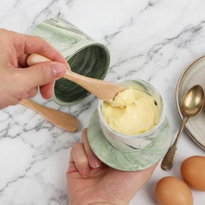 Puede incluir: Una mantequera con dise&ntilde;o de m&aacute;rmol verde y blanco con un cuchillo de mantequilla de madera. La mantequilla en el interior es amarilla. Tambi&eacute;n se ven un segundo plato y una cuchara.