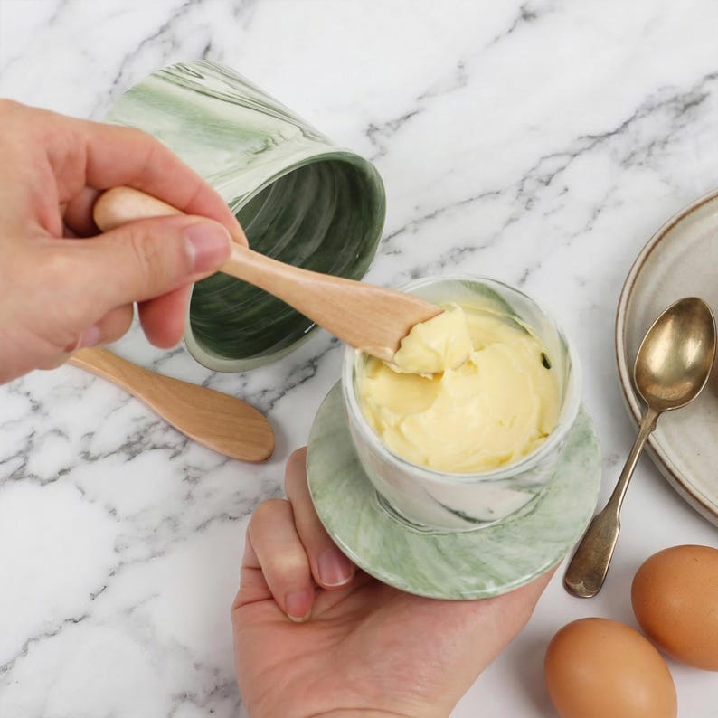 Puede incluir: Una mantequera con dise&ntilde;o de m&aacute;rmol verde y blanco con un cuchillo de mantequilla de madera. La mantequilla en el interior es amarilla. Tambi&eacute;n se ven un segundo plato y una cuchara.