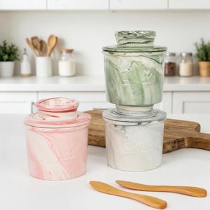 Puede incluir: Tres mantequilleras de cer&aacute;mica con tapas en patrones de m&aacute;rmol rosa, verde y blanco. Dos cuchillos de mantequilla de madera est&aacute;n en primer plano. Los platos son cil&iacute;ndricos con tapas a juego.