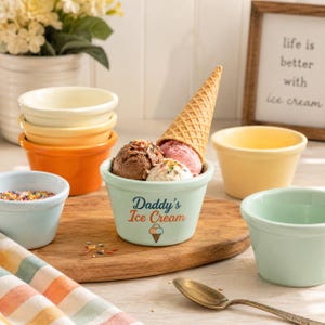 May include: A light green ceramic bowl with the text Daddy's Ice Cream and a small ice cream cone graphic. The bowl contains three scoops of ice cream topped with a waffle cone. Surrounding it are several empty colorful bowls, a spoon, and a cloth.