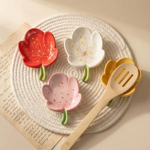Pode incluir: Quatro descansos de colher de cerâmica em forma de flor em vermelho, branco, rosa e amarelo. Cada um tem um caule verde e um design floral. Uma colher de madeira descansa no amarelo. O descanso de colher vermelho tem o nome "Bella".