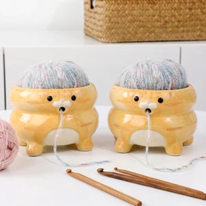 以下が含まれることがあります： オレンジ色の猫の形をしたセラミック製の毛糸入れが2つあり、それぞれに毛糸玉が入っています。ボウルには黒い目があり、糸を通すための小さな穴があります。かぎ針編みのフックが見えます。