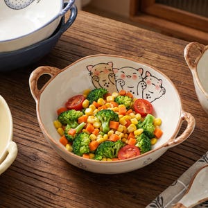 Peut inclure: Un bol en c&eacute;ramique blanc avec deux anses marron, rempli de l&eacute;gumes. Le bol est d&eacute;cor&eacute; de trois visages de chats de dessin anim&eacute;. Les l&eacute;gumes comprennent du brocoli, du ma&iuml;s, des carottes et des tomates. Le bol est sur une surface en bois.