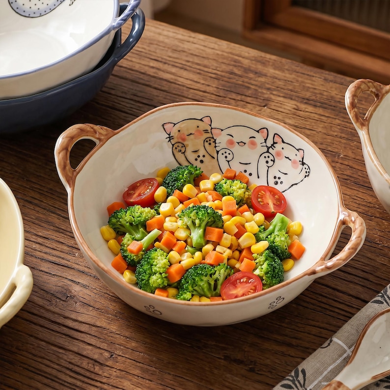 Peut inclure: Un bol en c&eacute;ramique blanc avec deux anses marron, rempli de l&eacute;gumes. Le bol est d&eacute;cor&eacute; de trois visages de chats de dessin anim&eacute;. Les l&eacute;gumes comprennent du brocoli, du ma&iuml;s, des carottes et des tomates. Le bol est sur une surface en bois.