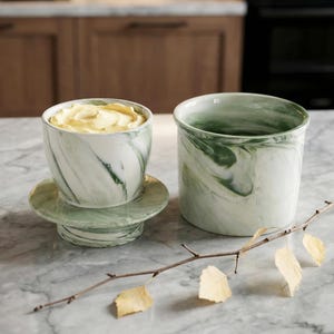 May include: Two ceramic butter dishes with a green and white marbled design. One dish is open, filled with yellow butter, and sits on a matching saucer. The other is a cylindrical container. A decorative branch with yellow leaves rests nearby.