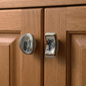 May include: Two ceramic cabinet knobs with a floral design in blue and gold. One knob is round, the other is rectangular. Both are mounted on a light brown wooden cabinet door.
