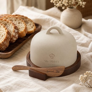 May include: A cream-coloured ceramic butter keeper with a dome lid featuring a handle, a chef hat icon, and the text "The Miller's EST 2026". It sits on a dark wood base with a matching wooden spreader knife engraved with the text "Spread Joy".