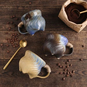May include: Three ceramic coffee mugs with unique, wavy designs in shades of blue, brown, and beige. Each mug has a rope-wrapped handle. A gold spoon and coffee beans are scattered on the wooden surface, along with a bag of ground coffee.