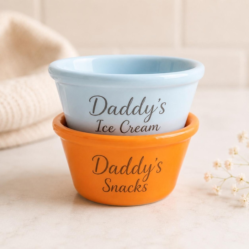 Pu&ograve; includere: Una ciotola in ceramica azzurra con la scritta Daddy's Ice Cream impilata all'interno di una ciotola in ceramica arancione con la scritta Daddy's Snacks. Entrambe le ciotole presentano una finitura liscia e lucida e un bordo arrotondato.