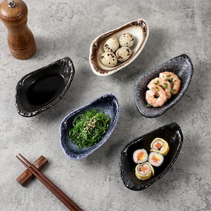 Pode incluir: Vista aérea de uma variedade de pequenos pratos de servir com formatos únicos. Os pratos contêm molho de soja, ovos de codorna, salada de algas, camarões e rolinhos de sushi. Um moedor de pimenta de madeira e pauzinhos também são visíveis.