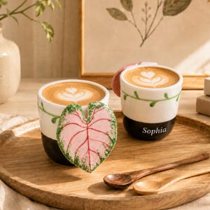Op de afbeelding: Twee keramische mokken gevuld met koffie en latte art. Eén mok heeft een groot, driedimensionaal roze en groen hartvormig blad aan de zijkant. De andere mok heeft een groen rankpatroon en de naam Sophia in witte letters gedrukt.
