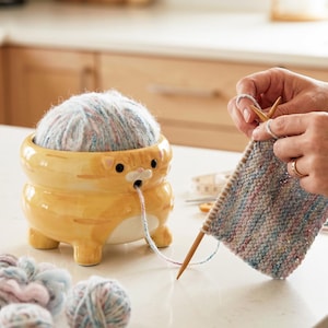 Könnte beinhalten: Ein gelb-oranges Keramik-Garnschalen-Katze mit einem Garnknäuel. Der Faden wird durch den Mund der Katze geführt. Stricknadeln und ein Strickstück sind im Hintergrund zu sehen. Das Garn ist mehrfarbig.
