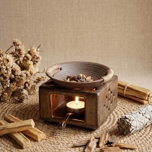 May include: A brown ceramic essential oil diffuser with a lit candle inside. The diffuser has a round bowl on top with wood pieces. Bundles of wood sticks and sage are arranged around the diffuser on a woven mat.