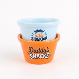 Pu&ograve; includere: Una pila di due ciotole in ceramica. La ciotola superiore &egrave; azzurra con un grafico a baffi blu scuro e il testo little SIDEKICK in arancione e blu scuro. La ciotola inferiore &egrave; arancione con il testo Daddy's SNACKS in blu scuro e bianco.