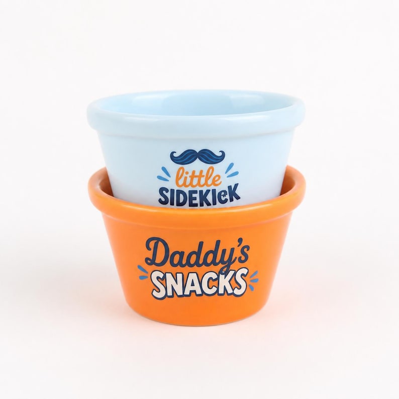 Pu&ograve; includere: Una pila di due ciotole in ceramica. La ciotola superiore &egrave; azzurra con un grafico a baffi blu scuro e il testo little SIDEKICK in arancione e blu scuro. La ciotola inferiore &egrave; arancione con il testo Daddy's SNACKS in blu scuro e bianco.
