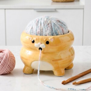 以下が含まれることがあります： 猫の形をした陶器製の毛糸入れで、中に毛糸玉が入っています。オレンジ色のボウルには黒い目があり、毛糸を通すための小さな穴があります。ピンク色の毛糸玉と2本の編み棒も写っています。