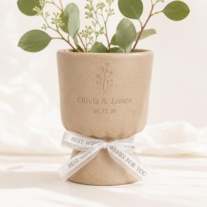 Può includere: Un vaso in ceramica beige con base scanalata e un nastro bianco legato attorno al centro. Il nastro riporta la scritta BEST WISHES FOR YOU. Il vaso è inciso con un rametto floreale, i nomi Olivia & James e la data 15.06.26.