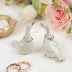 Peut inclure: Deux porte-anneaux en forme d'oie en céramique vert pâle, avec des becs dorés. Chaque oie porte une bague, une en or et une en argent, sur son cou. L'arrière-plan comprend des fleurs et un cadre décoratif, créant une présentation élégante.