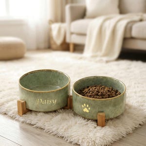 May include: Two green ceramic pet bowls with a gold rim and wooden stands. One bowl has the name "Daisy" in gold lettering, the other has a gold paw print and is filled with pet food. Bowls are on a fluffy white rug.